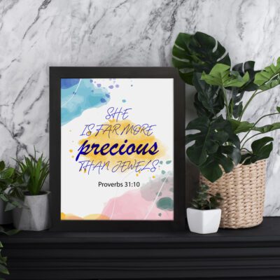 She Is Far More Precious Than Jewels Proverbs 31:10 Bible wall art with colorful watercolor splashes, blue and gold text. displayed on the table in a black frame