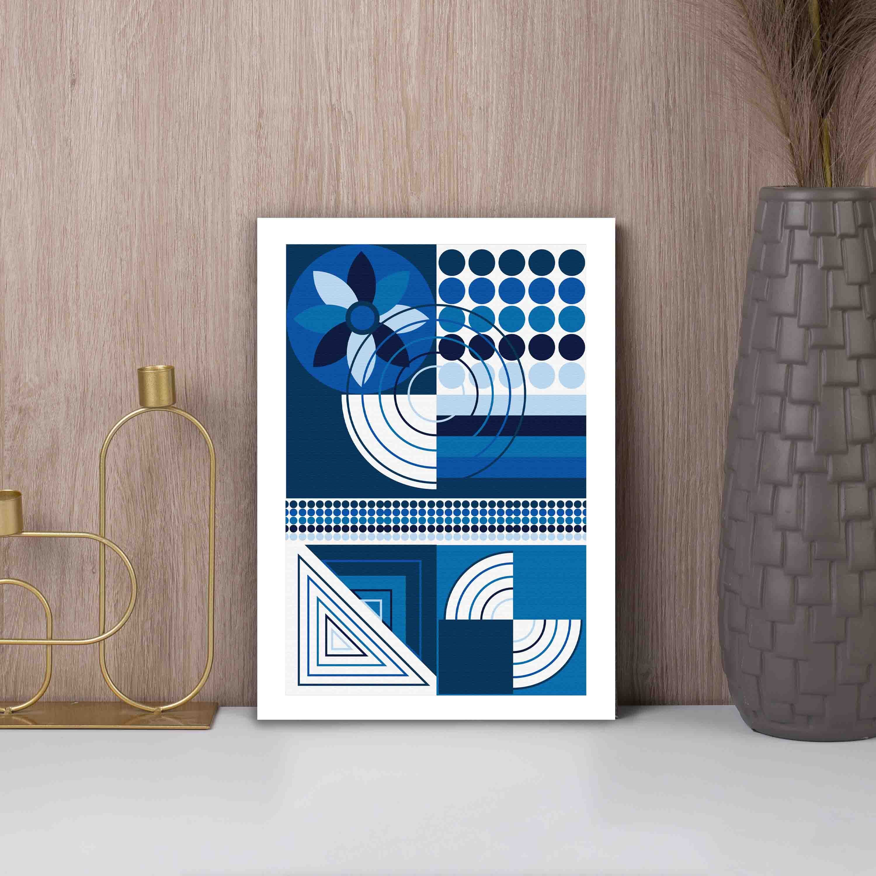 Abstract wall art with geometric shapes in blue, white, and black, framed in black, displayed on a light gray wall in a cozy setting.