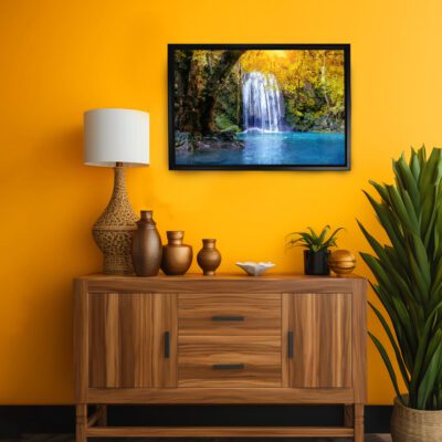 Waterfall wall art with lush greenery and a vibrant blue water body, framed and set against a bold yellow-orange background. displayed on the wall in a black frame