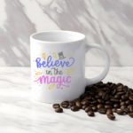 White coffee mug with Believe in the Magic in blue, black, and pink, decorated with a magician's hat and colorful elements. A microwave-friendly espresso cup.