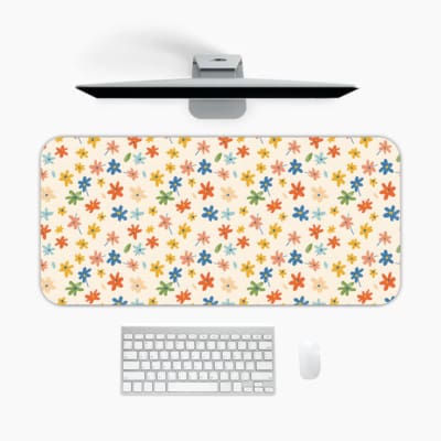 Colorful star-like flowers on a light pink background desk pad by Infidu, featuring vibrant red, blue, orange, green, and pink floral patterns. A desk pad under the computer