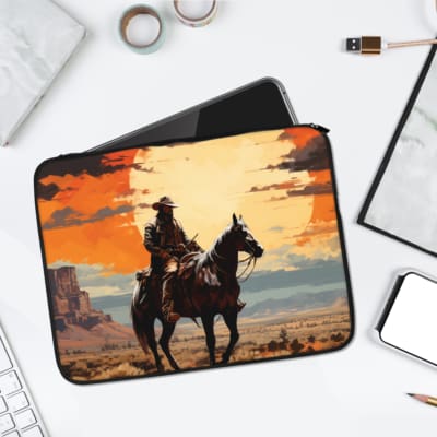 Laptop sleeve featuring a design of a cowboy riding a horse through a desert landscape during a sunset with orange, yellow, and blue tones. laptop sleeve kept on a plain white background