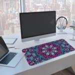 Infidu Colorful Mandala Design Desk Pad with intricate patterns in pink, blue, purple, and teal, plus white and black accents. A computer and laptop next to the desk pad