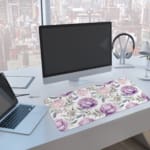 Infidu Purple Floral Design Desk Pad with large purple flowers and soft green leaves on a light, neutral background. A computer and laptop are next to the desk pad