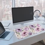 Infidu Flowers Design Desk Pad with green leaves and large purple and pink flowers on a light, neutral background. A computer and laptop are next to the desk pad