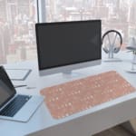 Infidu Beige Desk Mat with a subtle pattern of small shapes or icons on a soft beige background. A computer and laptop are next to the desk pad