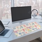 Desk pad with childlike drawings of flowers, stick figures, and hearts in pastel blue, pink, and yellow. A computer and laptop are next to the desk pad