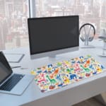 Bright desk mat featuring colorful animals, plants, and abstract shapes on a white background. A computer and laptop are next to the desk pad