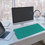 Desk pad with a turquoise green background and a subtle pattern. A computer and laptop are next to the desk pad