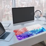 Desk pad with a gradient color design transitioning from red to blue and green, featuring abstract wavy patterns. A computer and laptop are next to the desk pad