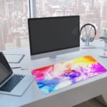 Infidu Vibrant Watercolor Design Desk Pad with abstract, colorful swirls of pink, orange, yellow, blue, and purple. A computer and laptop are next to the desk pad