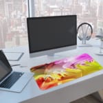 Infidu Colorful Abstract Wavy Lines Desk Pad featuring vibrant, flowing lines in pink, purple, yellow, orange, and green. A computer and laptop are next to the desk pad
