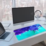 Infidu Abstract Desk Pad with blue, purple, and green colors, featuring sharp and flowing lines on a light background. A computer and laptop are next to the desk pad