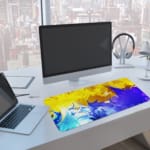 Infidu Abstract Desk Pad with yellow, blue, and green colors in flowing patterns on a vibrant background. A computer and laptop are next to the desk pad