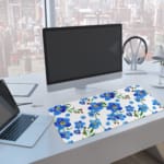 Desk pad with blue flowers, yellow centers, and green leaves on a white background. A computer and laptop are next to the desk pad