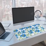 Desk pad with light blue flowers, yellow centers, and green leaves on a white background. A computer and laptop are next to the desk pad