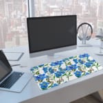 Blue floral desk pad with green leaves on a white background. A computer and laptop are next to the desk pad