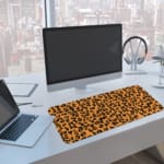 Infidu Leopard Print Design Desk Pad with black spots on a yellow background, simulating a leopard's natural fur. A computer and laptop are next to the desk pad