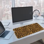 Infidu Leopard Print Desk Pad features a leopard print pattern with a brown background and black spots, resembling natural leopard fur. A computer and laptop are next to the desk pad