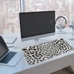 Infidu White Desk Pad with black leopard print spots on a white background, featuring a bold and untamed animal print design. A computer and laptop are next to the desk pad