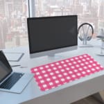 Pink and white checkered desk pad with a grid-like pattern on a white background. A computer and laptop are next to the desk pad