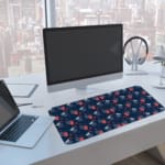 Infidu Red Strawberry and Blue Leaf Desk Mat with a dark blue background and a vibrant pattern of red strawberries and blue leaves. A computer and laptop are next to the desk pad