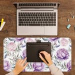 Infidu Purple Floral Design Desk Pad with large purple flowers and soft green leaves on a light, neutral background. A writing pad on top of the desk pad