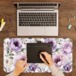 Infidu Flowers Design Desk Pad with green leaves and large purple and pink flowers on a light, neutral background. A writing pad on top of the desk pad