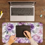 Infidu Floral Desk Pad featuring purple and light pink flowers with green and grayish leaves on a light white background. A writing pad on top of the desk pad