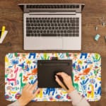 Bright desk mat featuring colorful animals, plants, and abstract shapes on a white background. A writing pad on top of the desk pad