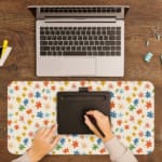 Colorful star-like flowers on a light pink background desk pad by Infidu, featuring vibrant red, blue, orange, green, and pink floral patterns. A writing pad on top of the desk pad