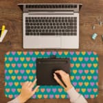 Infidu desk pad featuring colorful hearts in pink, yellow, and light purple on a teal background. A writing pad on top of the desk pad