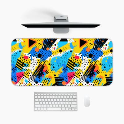 Infidu Colorful Abstract Desk Pad featuring playful geometric shapes in yellow, blue, pink, black, and turquoise on a white background. A desk pad under the computer