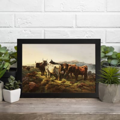 Vintage wall art of cows grazing in a serene countryside landscape with earthy tones and distant mountains. displayed on the table in a black frame