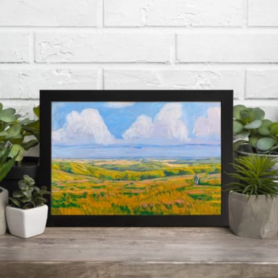 Infidu vintage wall art featuring a serene pastoral landscape with rolling hills, open fields, and soft clouds in the sky. displayed on the wall art in a black frame