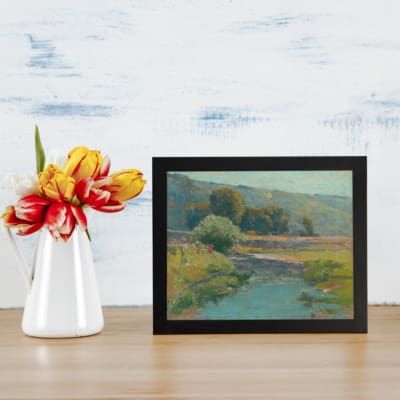 Infidu Vintage Wall Art featuring a tranquil river, rolling hills, and a peaceful natural landscape with soft, muted colors. displayed on the table in a black frame