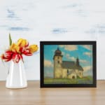 Vintage wall art of a rustic church in a calm countryside with a blue sky, white clouds, green grass, and touches of purple wildflowers. displayed on the wall
