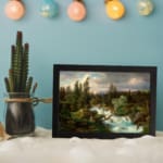 Vintage river flowing through a dense forest, with green trees and soft blue sky, framed in black. displayed on the wall