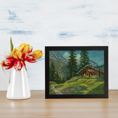 Vintage wall art of a countryside cabin surrounded by trees, hills, and a winding path with mountains and blue sky in the background. displayed on the table in a black frame