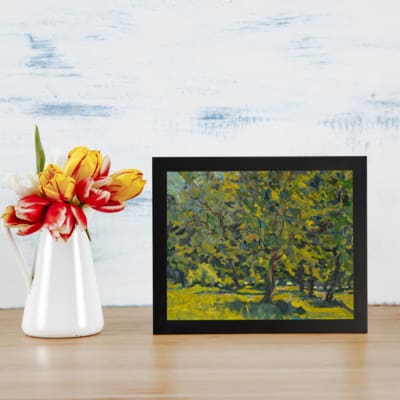 Vintage wall art with forest trees, lush foliage, and sunlight filtering through leaves in green and yellow tones. displayed on the table in a black frame