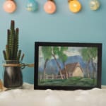 Vintage wall art of a countryside cottage with green trees and soft blue mountains. displayed on the wall