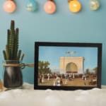 Vintage wall art of a 19th-century crowd in front of a grand white arch with flags, blue sky, and trees, framed in black. displayed on the wall
