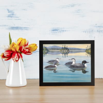 Vintage wall art of ducks swimming on a calm lake with trees and mountains in the background. Peaceful and timeless design. displayed on the table in a black frame