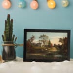 Infidu vintage wall art features a serene countryside scene with a charming house, tall trees, and a soft blue sky. displayed on the wall art in a black frame