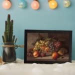 Rustic still-life painting of peaches, grapes, and pears in warm tones with a brown background and black frame. displayed on the wall