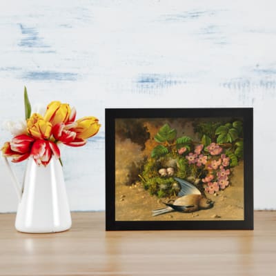 Infidu Vintage Wall Art featuring a bird nest with a bird perched near blooming flowers in serene and rustic nature-inspired design. displayed on the table in a black frame