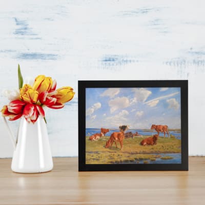 Vintage wall art featuring cows grazing in a green field near water under a blue sky with clouds, perfect for rustic and countryside decor. displayed on the table in a black frame