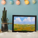 Infidu vintage wall art featuring a serene pastoral landscape with rolling hills, open fields, and soft clouds in the sky. displayed on the wall art in a black frame