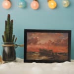 Vintage wall art of a wooden ship at sunset with fiery red, orange, and pink sky, calm sea, and small human figures near the shore. displayed on the wall