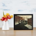 Vintage winter river scene with brick buildings and snowy rooftops by Infidu. displayed on the wall